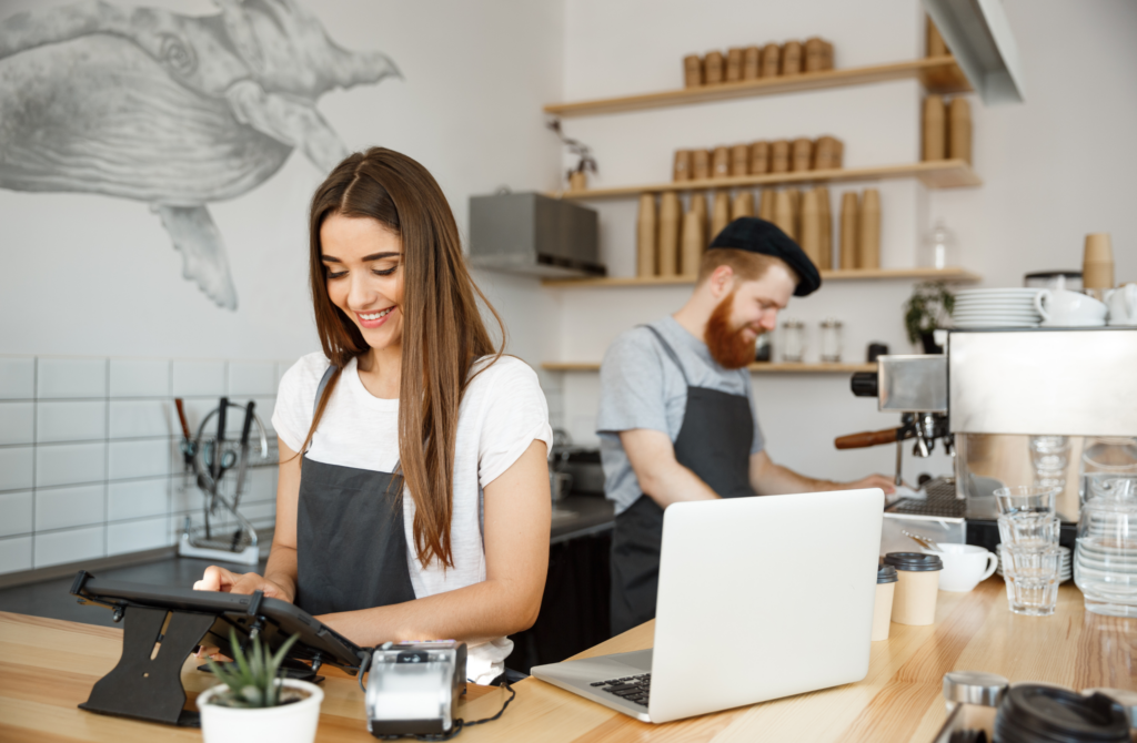 Woman in a white shirt and apron using a tablet at a wooden cafe counter, with a bearded barista in a black beret in the background.